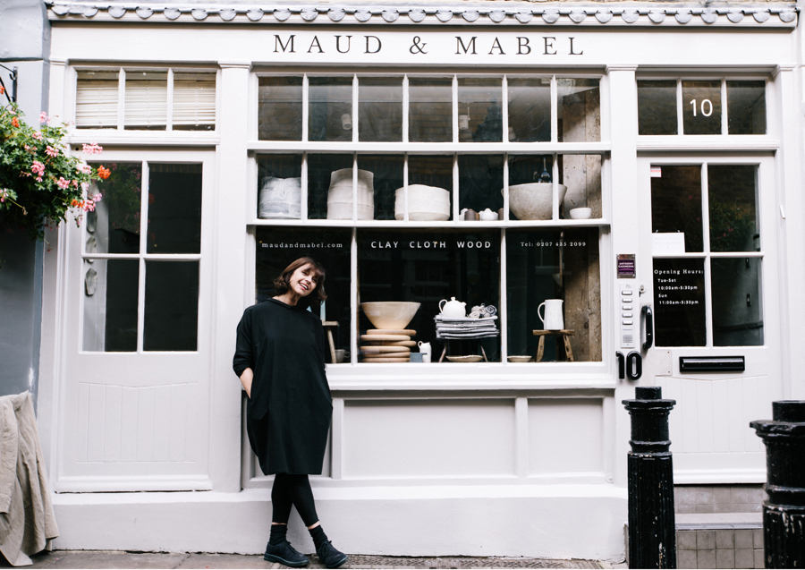 Woman with brown hair and black dress stands in front of simple shop front with ceramics in the windows. Maud & Mabel