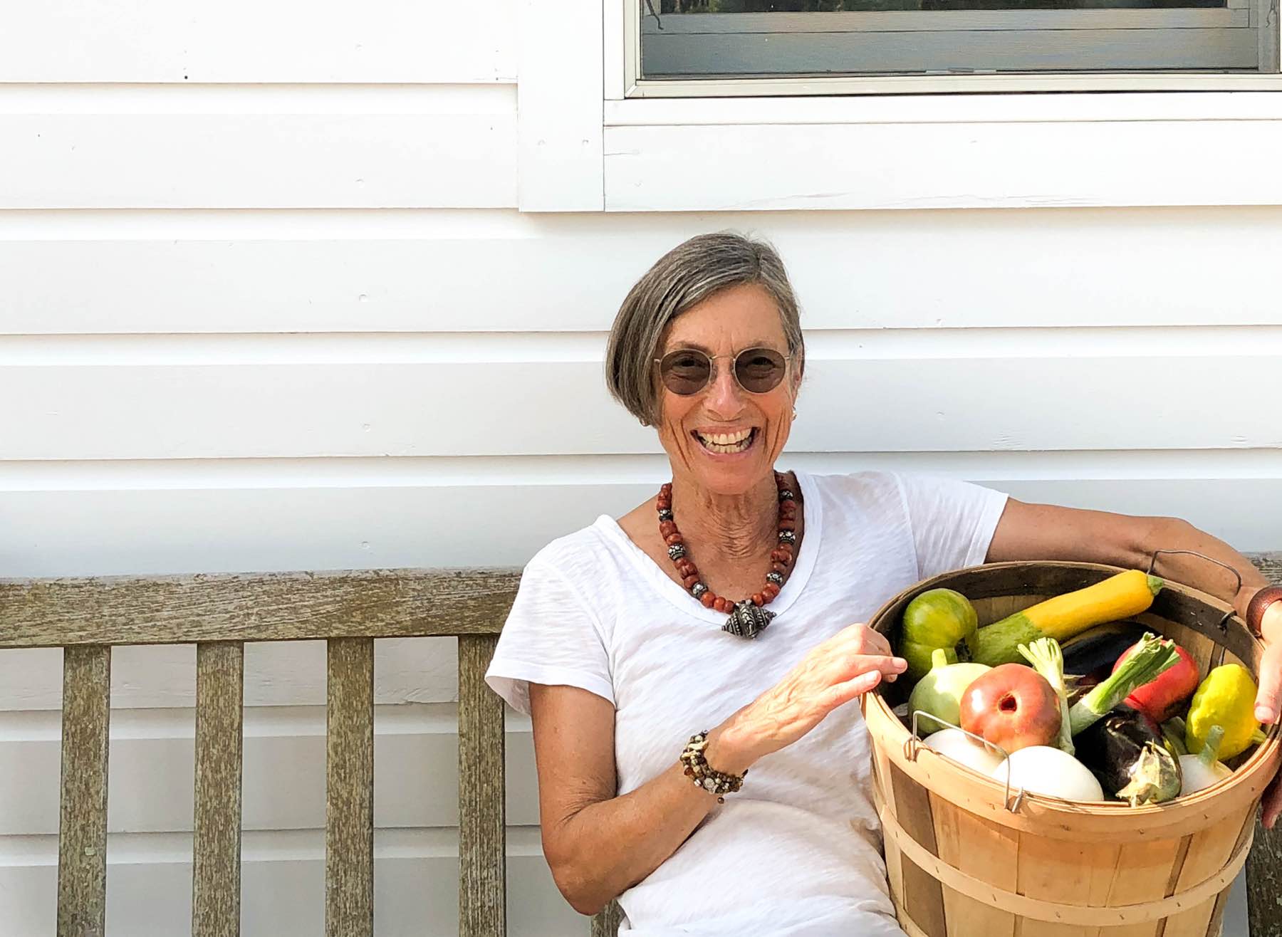 Woman on bench in front of white clapboard holding a basket of summer vegetables, White Gate Farm